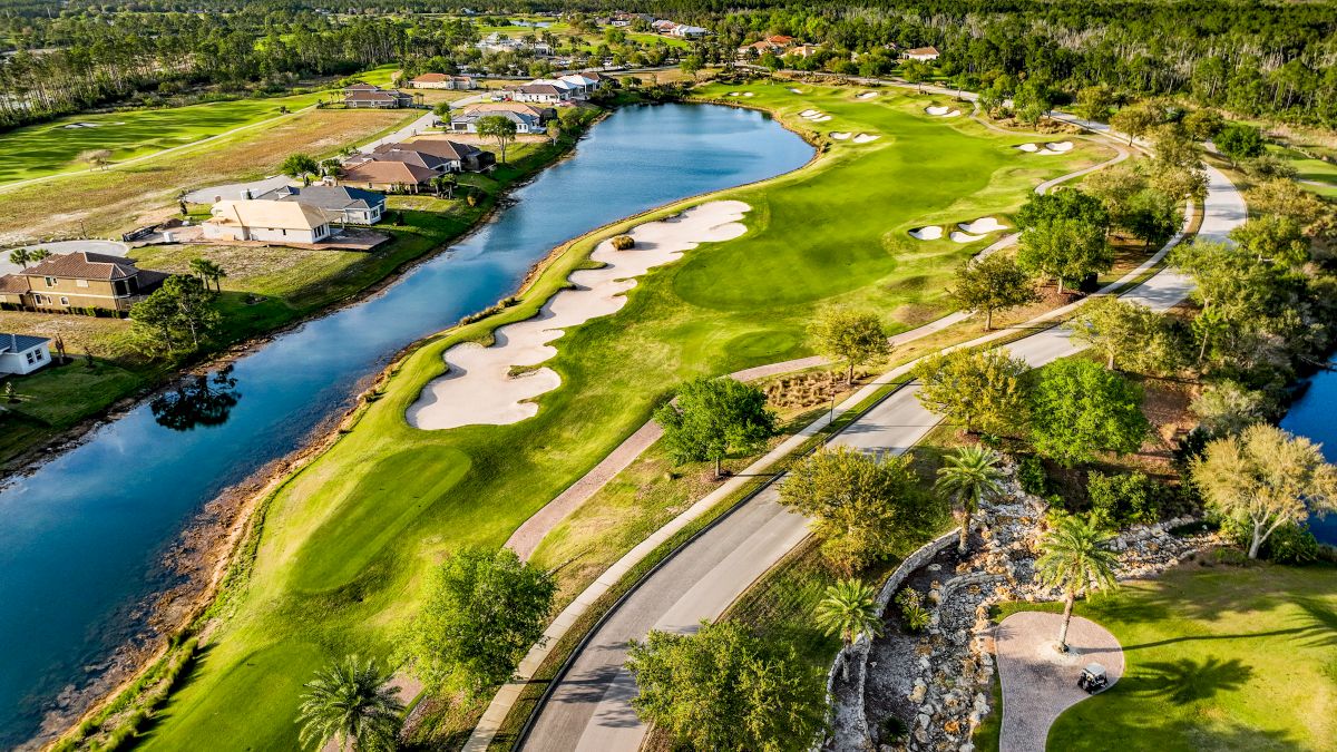Aerial view of a golf course with sand traps and a water hazard, bordered by houses and a tree-lined road.