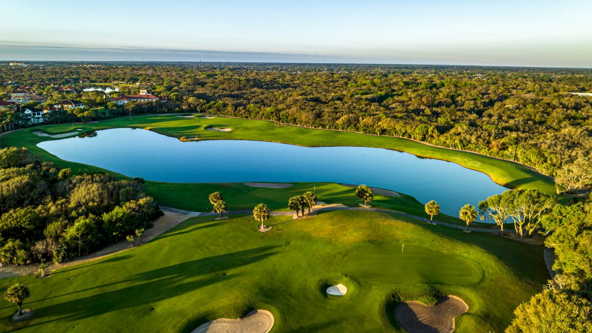 Aerial view of a golf course with a lake, green fairways, sand bunkers, and surrounding trees under a clear blue sky.