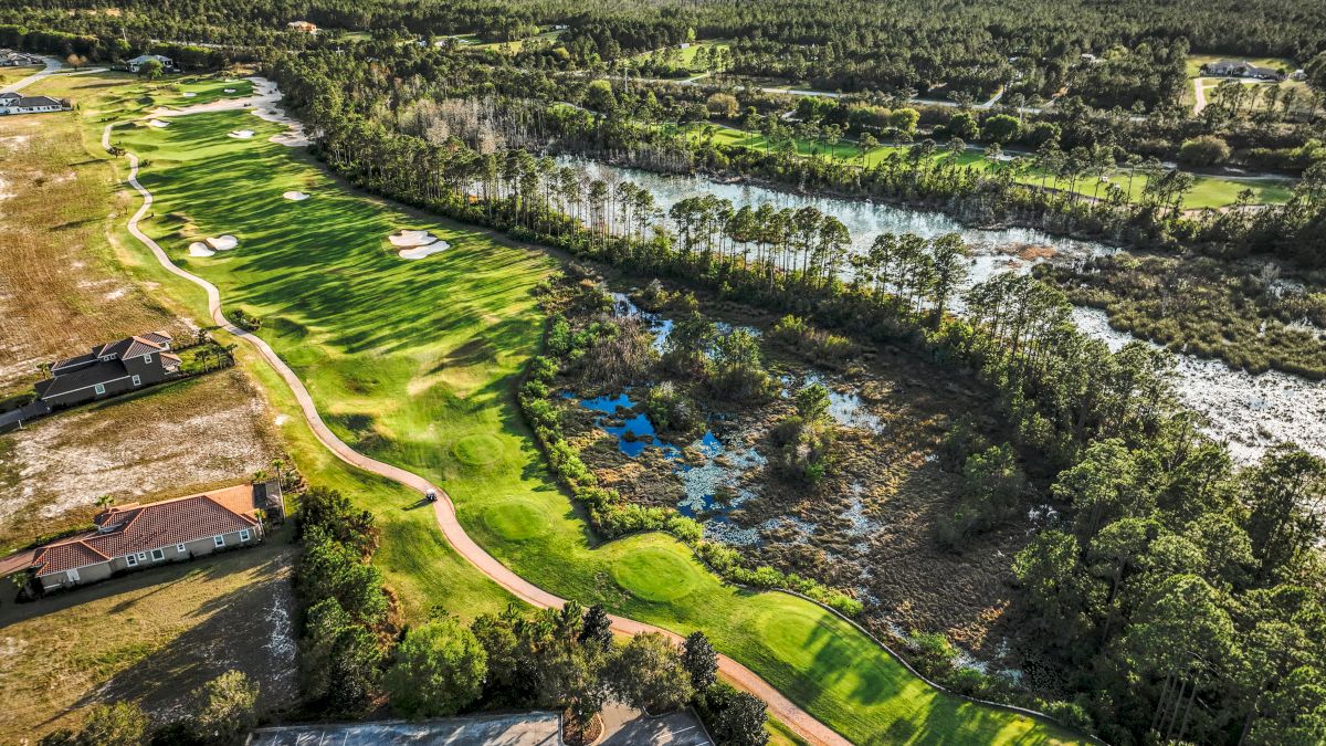 Aerial view of a lush golf course with green fairways, surrounded by trees and water bodies, adjacent to residential and parking areas.