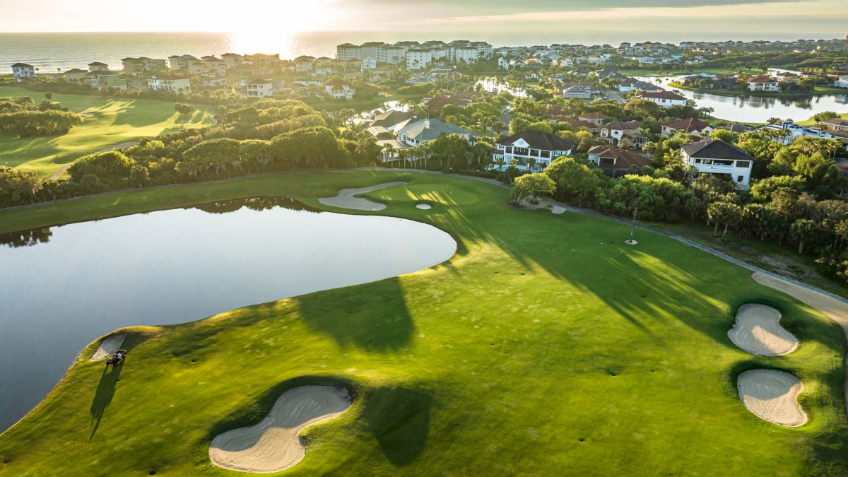 Aerial view of a golf course with sand bunkers and a water feature, surrounded by residential houses near the ocean, during sunset.