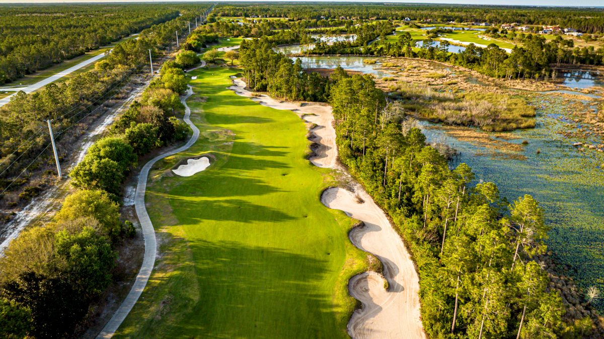 An aerial view of a golf course surrounded by trees and water features, with sand bunkers and pathways visible throughout the scene.