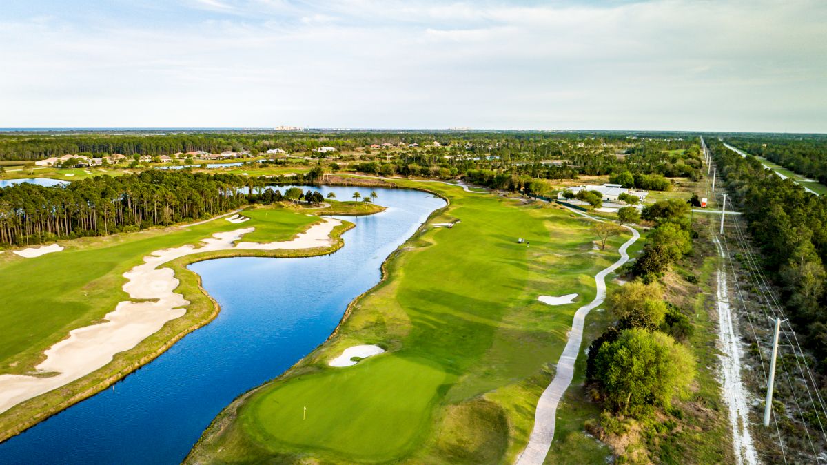 An aerial view of a golf course with lush greens, sand bunkers, a winding water body, and surrounding trees under a vast blue sky.