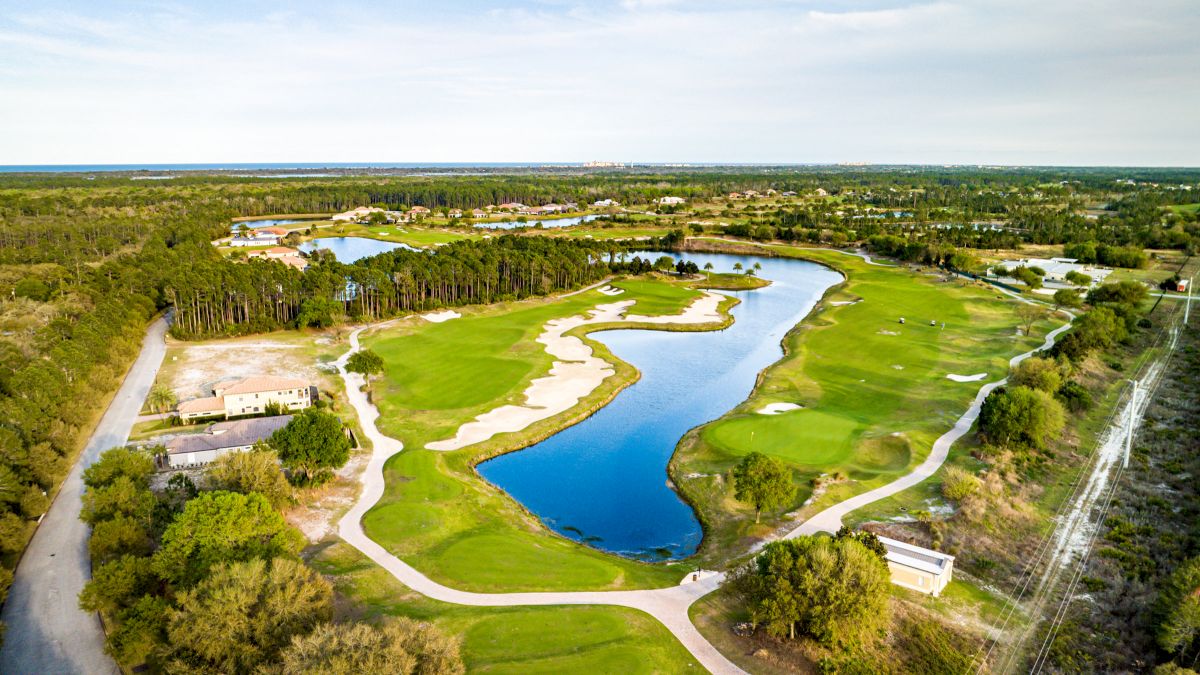 Aerial view of a green golf course with sand traps and a water hazard, surrounded by trees and a clear sky.