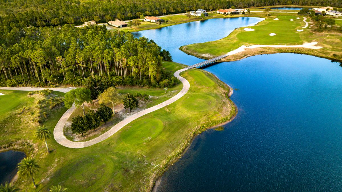 Aerial view of a golf course with a winding path, trees, a lake, and several buildings in the background.