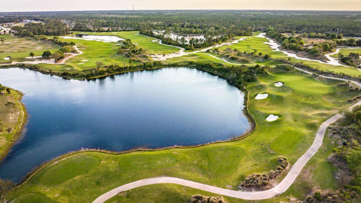 An aerial view of a golf course with a large pond, green fairways, sand bunkers, and winding paths surrounded by trees and a vast forest.
