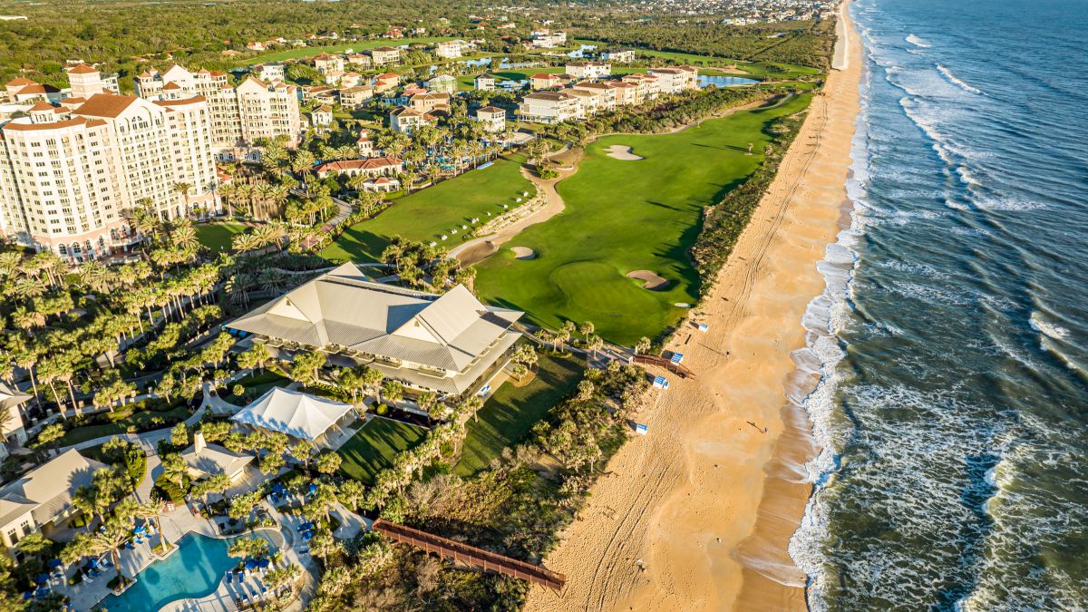 Aerial view of a beachfront resort with pools, a golf course, and the ocean. The coastline stretches into the horizon.
