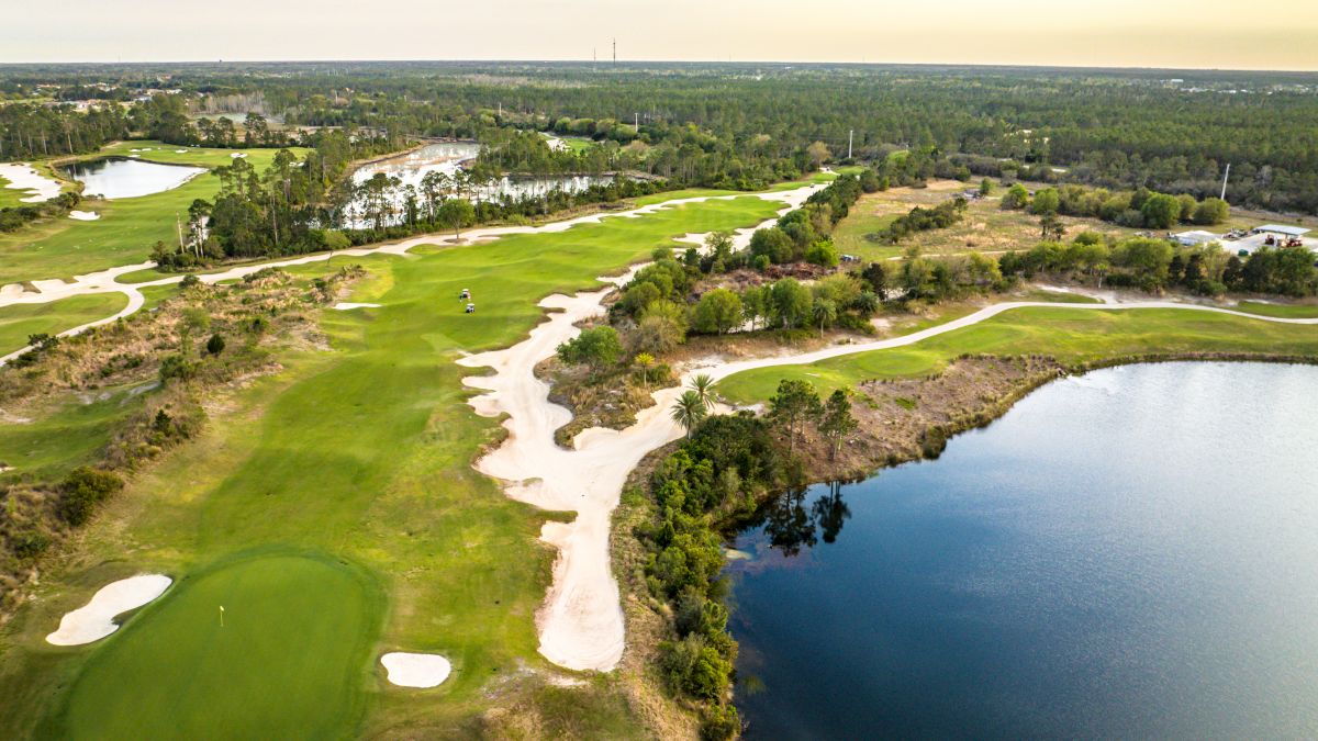 The image shows a scenic aerial view of a golf course with green fairways, sand traps, and a water body on the right side.