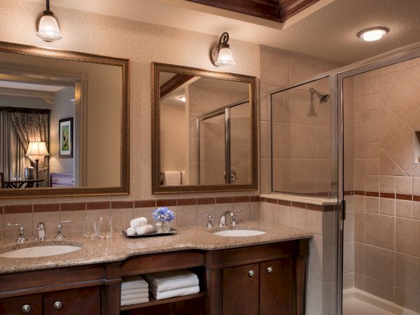This image shows a bathroom with two sinks, mirrors, and a shower. A neatly arranged cabinet with towels is below the sinks.