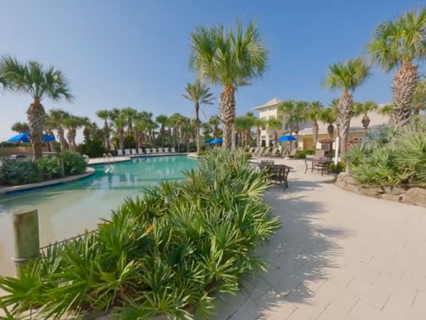 The image shows a scenic pool area surrounded by palm trees and lush greenery under a clear blue sky, creating a tropical atmosphere.