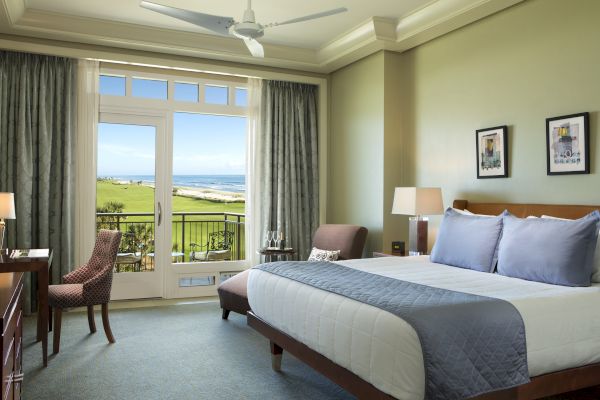 A well-appointed hotel room features a large bed, cozy chairs, a desk, and a balcony with a scenic view.