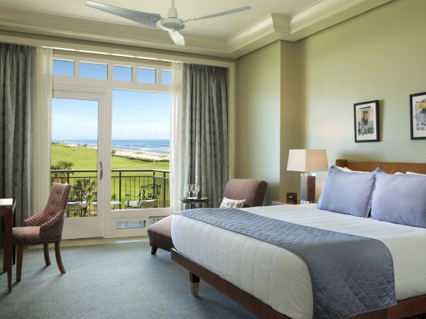 A well-appointed hotel room features a large bed, cozy chairs, a desk, and a balcony with a scenic view.