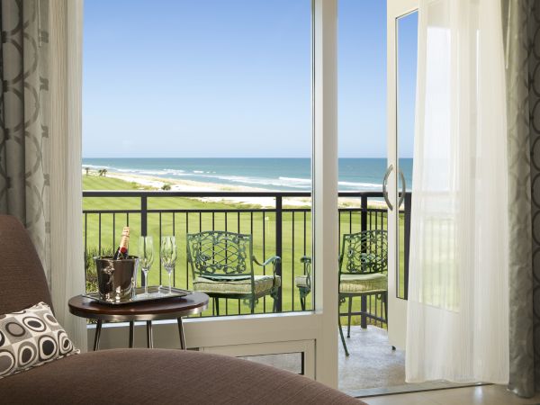 A cozy room with an open door to a balcony, offering a beach view. There's a small table with drinks and chairs outside.