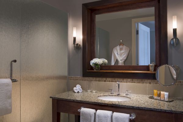 A bathroom with a granite countertop, large mirror, towels, bath products, and soft lighting. A robe hangs on the door in the background.