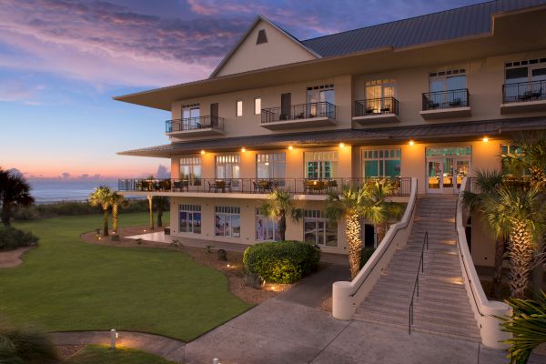 A large, well-lit building with balconies and stairs surrounded by greenery and a sunset sky, near a body of water, likely coastal.