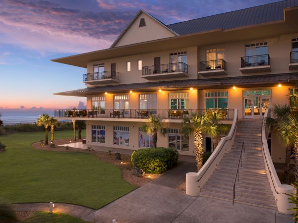 A large, well-lit building with balconies and stairs surrounded by greenery and a sunset sky, near a body of water, likely coastal.