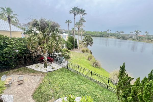 A lakeside backyard with a patio, palm trees, and greenery, overlooking a calm body of water under a cloudy sky.