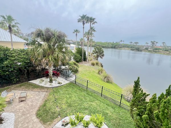 A lakeside backyard with a patio, palm trees, and greenery, overlooking a calm body of water under a cloudy sky.