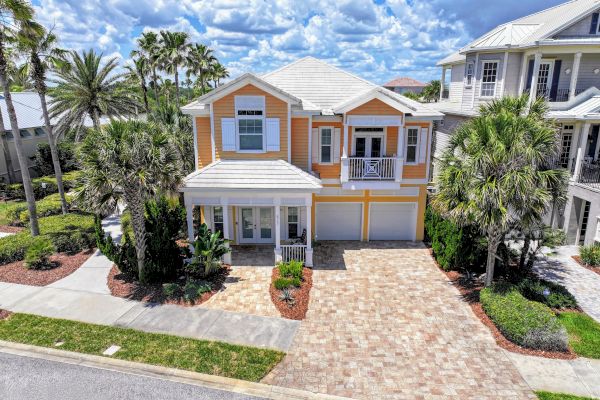 A two-story house with a warm color scheme, surrounded by palm trees and landscaped areas, with a paved driveway under a partly cloudy sky.