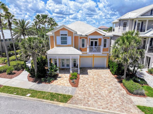A two-story house with a warm color scheme, surrounded by palm trees and landscaped areas, with a paved driveway under a partly cloudy sky.