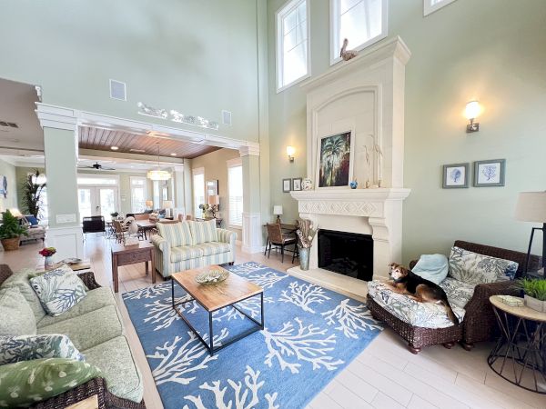 A spacious living room with high ceilings, comfy furniture, a fireplace, and large windows. A dog relaxes on a chair.