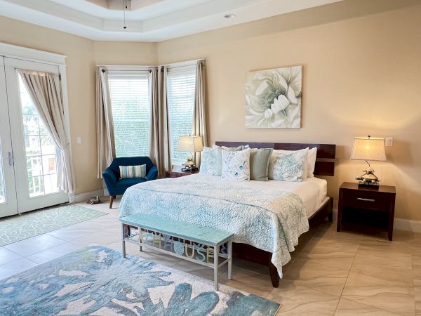 A spacious bedroom with a large bed, two bedside tables with lamps, and a sitting area by windows with curtains and patterned decor.