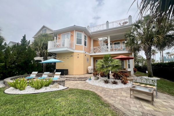 A two-story house with balconies, patio, lounge chairs, umbrellas, palm trees, and manicured garden on a cloudy day ends the sentence.