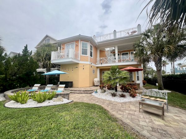 A two-story house with balconies, patio, lounge chairs, umbrellas, palm trees, and manicured garden on a cloudy day ends the sentence.
