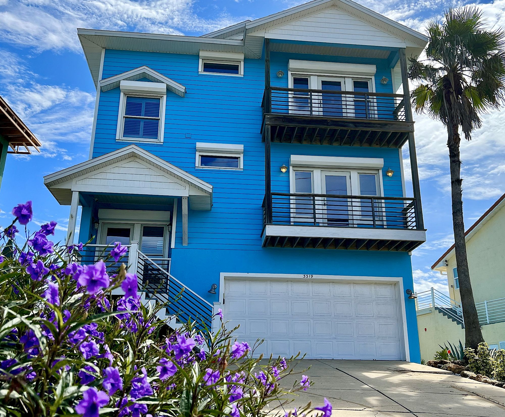 A vibrant blue three-story house with balconies, a white garage, surrounded by purple flowers and a palm tree under a bright sky.