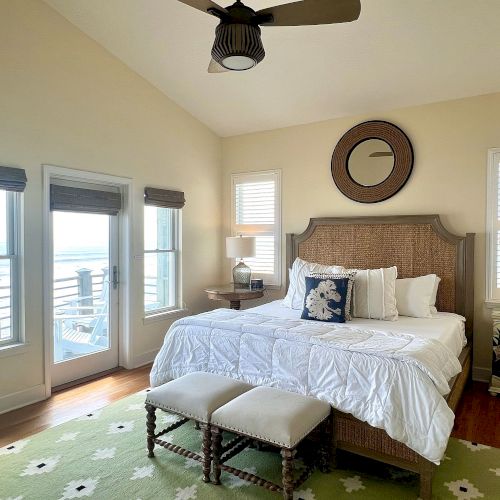 A bedroom with a large bed, decorative pillows, a green rug, two benches, and a window view of a balcony with ocean scenery.