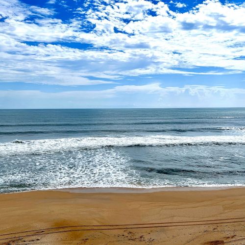 A sandy beach with gentle waves meets the ocean under a partly cloudy blue sky.