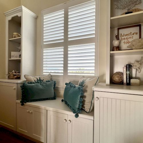 A cozy nook with white cabinets, teal pillows, and decorative shelves featuring books and coastal accents, next to a window with shutters.