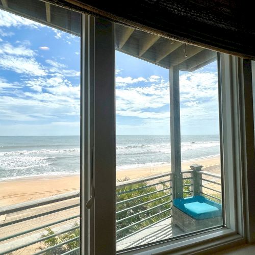 A view from inside showing a beach, ocean, and partly cloudy sky through large windows with a blue cushion on a railing.