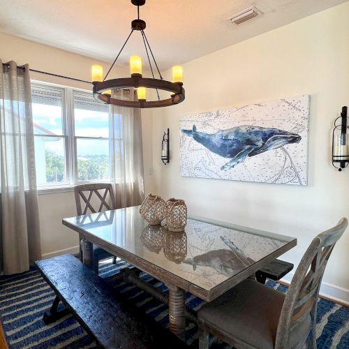 A dining room with a large table, chandelier, whale artwork, striped rug, and decorative jars, next to a window with curtains.