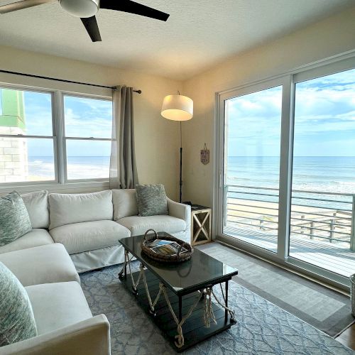 A cozy living room features a white L-shaped sofa, large windows, and a view of the ocean. Decor includes a rug, table, and floor lamp.