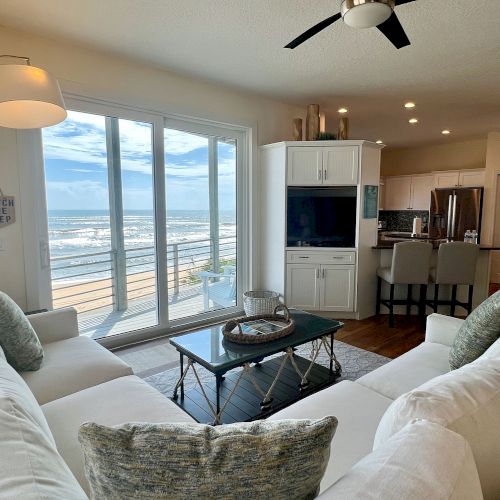 A modern living room with ocean view, white sofas, a coffee table, large windows, kitchen with bar stools, and ceiling fans.
