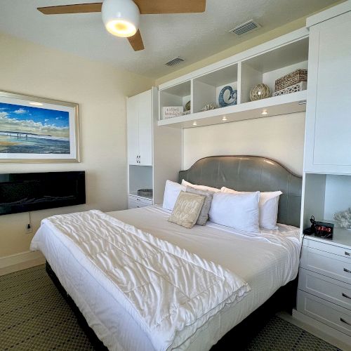 A cozy bedroom with a made-up bed, ceiling fan, wall art, TV, storage cabinets, and decorative items on shelves, creating a peaceful ambiance.