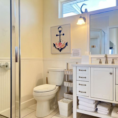 The image shows a clean bathroom with a glass shower, toilet, and a white vanity. Above the toilet is a nautical-themed anchor artwork.