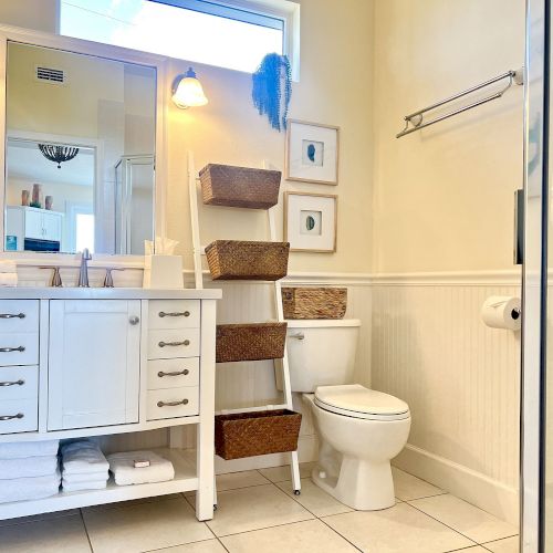 A bathroom with a white vanity, mirror, toilet, wicker basket shelves, towel rack, and tiled floor.