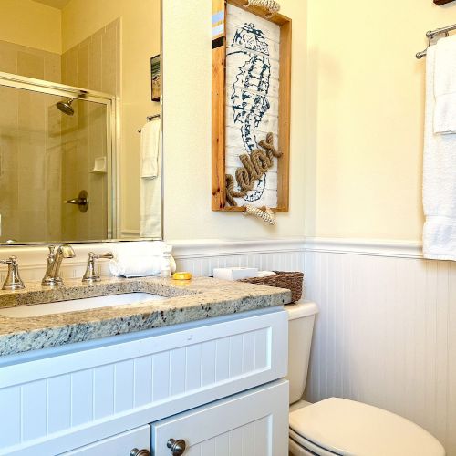 A bathroom with a granite countertop, sink, towel rack, toilet, and a shower with a glass door. Decor includes nautical wall art.