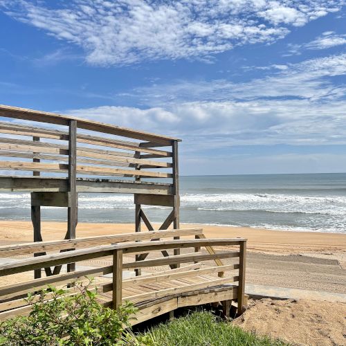A wooden boardwalk leading to a sandy beach with ocean waves under a partly cloudy blue sky.