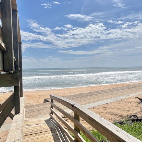 A sandy beach view with a wooden pathway leading to the ocean, under a partly cloudy blue sky and gentle waves.