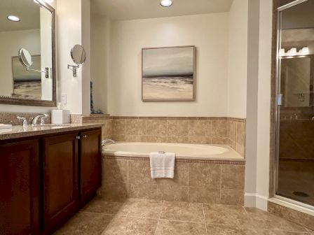 A bathroom with a bathtub, tiled walls, wooden cabinets, a mirror, and a shower. A painting hangs above the tub.