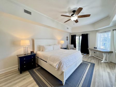 A cozy bedroom with a white bed, blue nightstand, lamps, ceiling fan, desk, and chair in a sunlit room with wooden flooring.