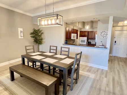 A dining area with a wooden table, chairs, and bench; kitchen in the background with cabinets, appliances, and a wall clock.