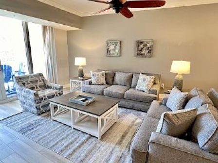 A modern living room with gray sofas, a patterned chair, a coffee table, lamps, and wall art, featuring a ceiling fan above.