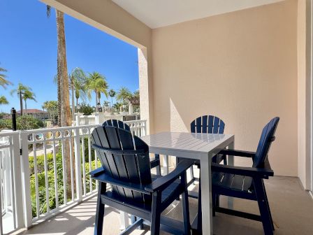 A balcony with a white table and blue chairs overlooks palm trees and a sunny outdoor scene.