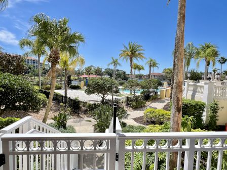 A sunny outdoor scene with palm trees, a pool area, and a garden walkway, bordered by a white fence.