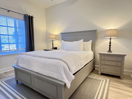 A bedroom with a large bed, white bedding, two matching lamps on nightstands, and a window with closed blinds and curtains.