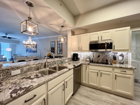 A modern kitchen with beige cabinets, granite countertops, metallic appliances, and elegant lighting fixtures. Open layout leads to the living area.