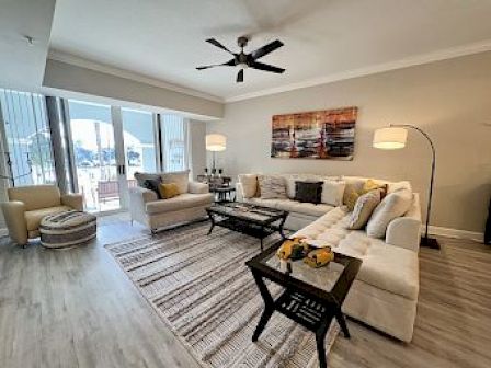 A modern living room with a beige sectional sofa, coffee table, floor lamp, ceiling fan, artwork, and large windows with a view.